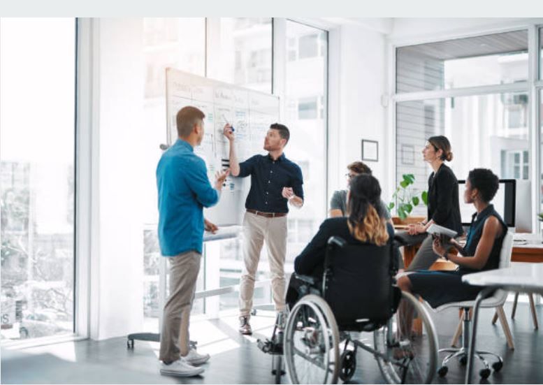 Picture of work colleagues at a white board including a person of color and a person using a wheelchair
